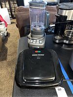 Cuisinart blender with clear plastic pitcher and several buttons on the metallic base, next to the George Foreman black electric grill with hinged lid, placed on a table in a living room setting.