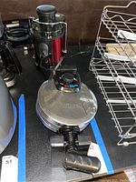 Photo showing a metallic round Bella waffle iron with a black handle and temperature dial alongside a black and red Enjapp electric juicer with clear plastic top