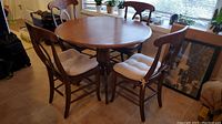 Full view of round wooden table with four matching chairs with white cushions placed around it, natural light from window