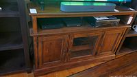 Front view of wood TV stand showing three doors, one with a glass etched panel, surface with scratches.
