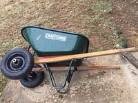 Side and top view of the green metal basin with wooden handles and black pneumatically inflated wheel showing rust, placed on concrete ground outdoors.