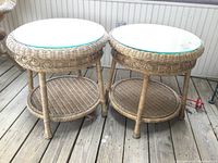 Pair of round wicker side tables with clear glass tops on wood flooring. Both tables have woven wicker lower shelves and legs.