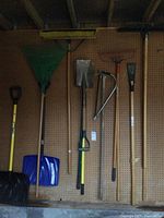 Photo of yard tools hanging on pegboard in shed showing snow shovels, leaf rake, digging shovel, broom, hand saw, and other shovels and rakes.