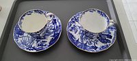 Two blue and white patterned cups on matching saucers seen from a slight angle on a gray tray.
