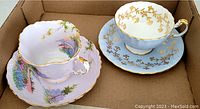 Overhead view of two Aynsley cups and saucers in a cardboard box, showing detailed lavender scenic design on one set and blue with gold leaf decoration on the other.