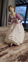 Front and side view of the Royal Doulton porcelain figurine featuring a woman in a white skirt and pink blouse holding a small purple flower.