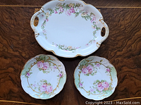 Overview of two small floral porcelain plates and one large serving tray with cutout handles, all hand painted in pink, green and gold.