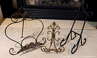 Three assorted decorative metal stands of different colors and ornamental designs photographed on tile floor.