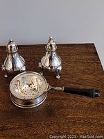 Pair of silver plated salt and pepper shakers with ornate footed bases and removable tops. Tea strainer with black handle resting in matching silver plated bowl.