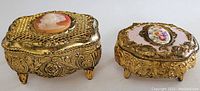 Two ornate vintage trinket boxes side by side, showing gold-tone metal relief detail, cameo and floral medallion tops