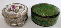 Two trinket boxes side by side: silver-tone metal with floral porcelain inset and green wooden box with gold leaf design.