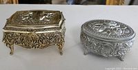 Two metal trinket boxes shown from front view with detailed ornate relief designs.