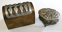 Photo showing two metal trinket boxes; a rectangular box with a brass base and silver cutout design lid, and a smaller heart-shaped embossed box.