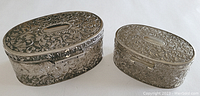 Two oval metal trinket boxes shown top side up, highlighting the larger and smaller box size difference, intricate floral and scrollwork embossing, and empty oval cartouche on the lid.