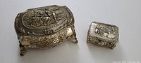 Two metal trinket boxes side by side, showing embossed romantic scenes on lids and intricate side detailing.