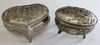 Photo showing metal trinket boxes, heart-shaped and oval with embossed designs, resting on white background.