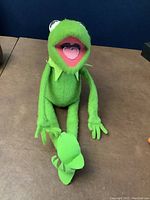 Front view of the Kermit the Frog plush showing the face, felt collar, and seated pose with extended legs.