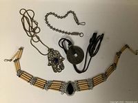 Photo showing three necklaces and one bracelet laid out on a flat surface. The bead bracelet features beige elongated beads and rhinestone details with a central black stone. Necklaces include a silver Hamsa hand pendant with blue stone, a ball chain with silver end caps, and a black cord with round pendant.