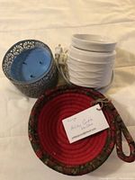 Photo showing a white ribbed electric aroma diffuser, a blue metal candle holder with open lattice design, and a red braided circular mat with a handle.