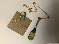 Photo showing a wooden paddle-shaped necklace pendant with green accents on a gold chain alongside a brass-colored bobby pin hair clip with a round decorative top showing a tree design and the words 'Love Life'. Both items are on a white background.
