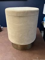 Front view of the plush round stool showing sesame fabric and antique brass base.