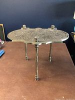 Side view of the small brass table showing its three slender legs and the detailed ornate tabletop