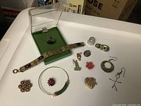 Photo showing jade cuff bracelet, black onyx cufflink or ring in case, various earrings, buttons including floral designs, thimble, and small figurines arranged on a white surface.