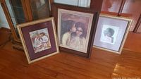 Photo showing three framed art pieces on wooden floor: two gold framed works with matting and one dark brown framed print with children.