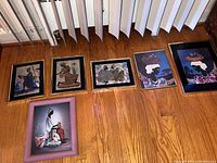 Photo showing five of the six framed artworks laid against a wall on a wooden floor. Art depicts stylized African and tribal women and scenes with black, gold, and pink frames.