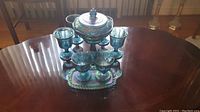 Photo showing the full set on a wooden table including the covered candy dish, four goblets, creamer, sugar bowl, and tray with iridescent glass finish.