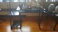 Full view of smoked glass computer desk with side shelves and black metal frame on hardwood floor.