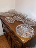 Pressed glass serving pieces arranged on wooden cabinet including platters and dishes with floral and starburst patterns