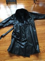 Full view of the laid flat black leather coat showcasing length, sleeves, front belt, and pockets on a hardwood floor.