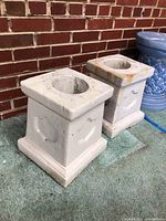 Two square concrete planters shown side by side next to brick wall and blue decorative pot in background. Both are off-white with some staining and decorative panels on sides.