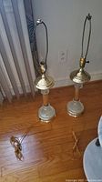 Photo showing two glass and metal table lamps with cords on a wooden floor near vertical blinds and an electrical outlet.