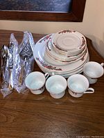 Fine china dinnerware and silver plated utensils arranged on wooden surface, showing four cups, various plates, and utensils in plastic wrapping.