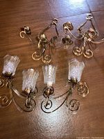 View of four metal wall sconces with gold-tone finish laid on wooden surface. Each has curved arm design and clear textured glass candle holders for candles.
