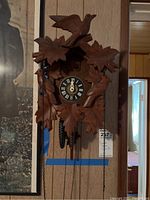 Full view of the carved wooden cuckoo clock mounted on a wall, showing the bird and leaf carvings, clock face, pinecone weights, and chains.