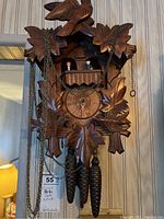 Front close-up view of the cuckoo clock showing detailed wood carvings and figurines on balcony with hanging pine cone weights
