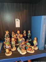 Wide shot of multiple Goebel Hummel porcelain figurines displayed on wooden shelf next to a Goebel Collectors Club binder.