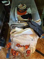 Photo of two Orioles caps, multiple white and black clothing items, and a white backpack with Orioles logo, with four clear glass mugs in front.