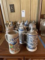 Front-facing view showing 11 beer steins and one large mug arranged on a wooden cabinet top, displaying various ceramic and metal lid styles.
