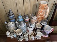 Photo showing the entire collection of 7 steins and 7 mugs arranged against a wall, highlighting the tallest tan stein with relief artwork and varied smaller steins and mugs in front.