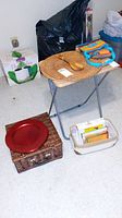 Overall view of lot items displayed on small table and floor, showing placemats, wood utensils, charger plate, picnic basket, and cookbooks