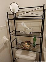 Black metal wire bathroom rack with 3 shelves placed above a toilet tank, featuring a box of tissues and small personal items.