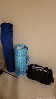 Photo showing all three items: folded blue canvas beach chair, rolled light blue quilted beach mat with pillow, and black canvas tote bag against wall.