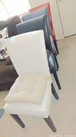 Four multi-color faux leather dining chairs arranged on carpet, showing cream chair in front with beige cushion and other chairs in navy blue, teal, and red behind.