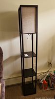 Front view of tall black plastic floor lamp with three square shelves and fabric-covered light diffuser at the top.