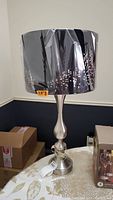 Full view of the lamp showing brushed silver toned metal base with a black lampshade decorated with a stenciled city skyline pattern, placed on a table surrounded by boxes.