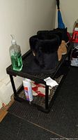 Black plastic shoe rack with two shelves shown loaded with various shoes and miscellaneous items including a bottle of hand sanitizer and a canister.